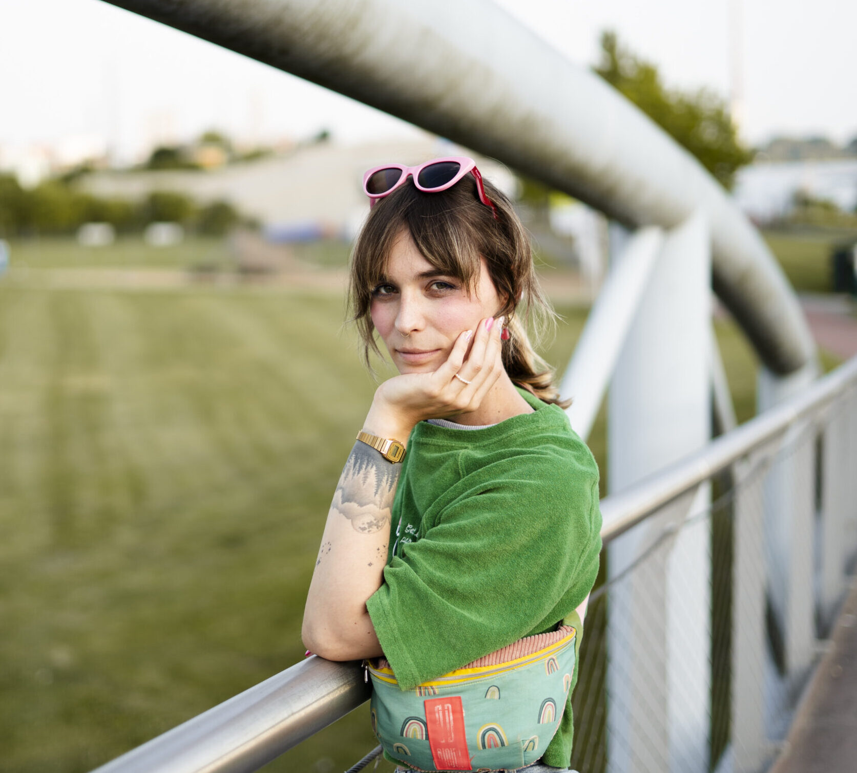 Nyke Slawik lehnt auf einem Metallgeländer im Freien, trägt grünes T-Shirt, rosa Sonnenbrille auf dem Kopf und eine bunte Bauchtasche.