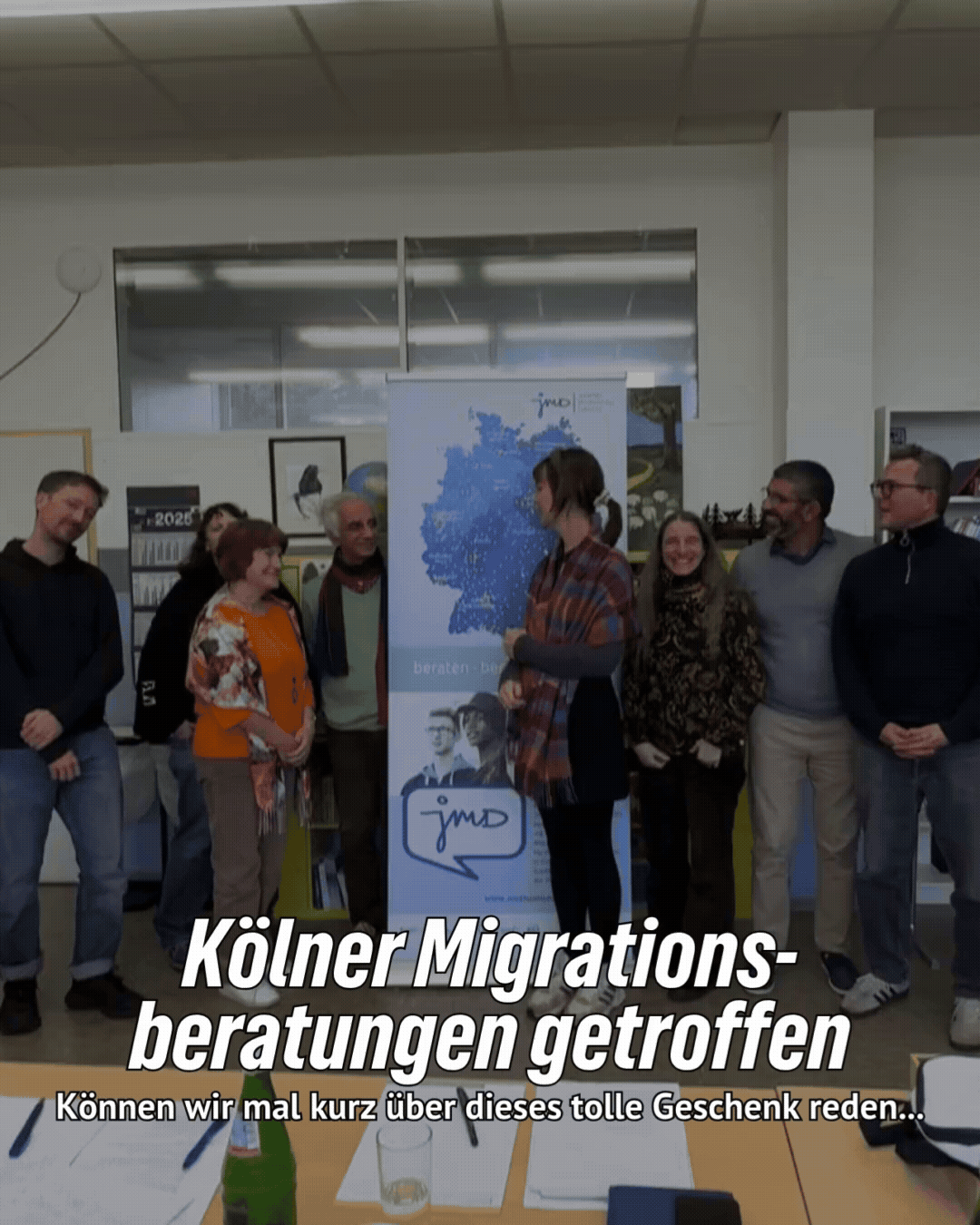 Acht Personen posen in einem Raum für ein Foto, vor einem Banner von jmd. Text: Kölner Migrationsberatungen getroffen - Können wir mal kurz über dieses tolle Geschenk reden..."